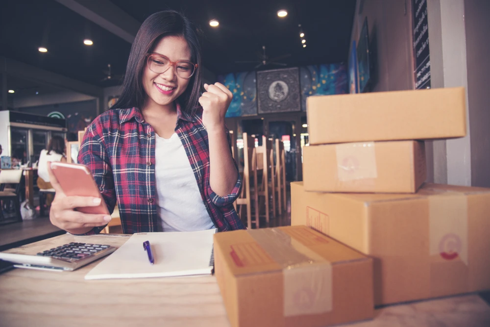 Woman smiling at her phone beside shipping boxes, representing ecommerce sales, online orders, and small business digital marketing.